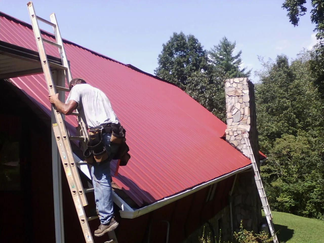 Roofer on a ladder beside a red metal roof and stone chimney — Soffit & Fascia Repair by Roofing Brothers in Tennyson